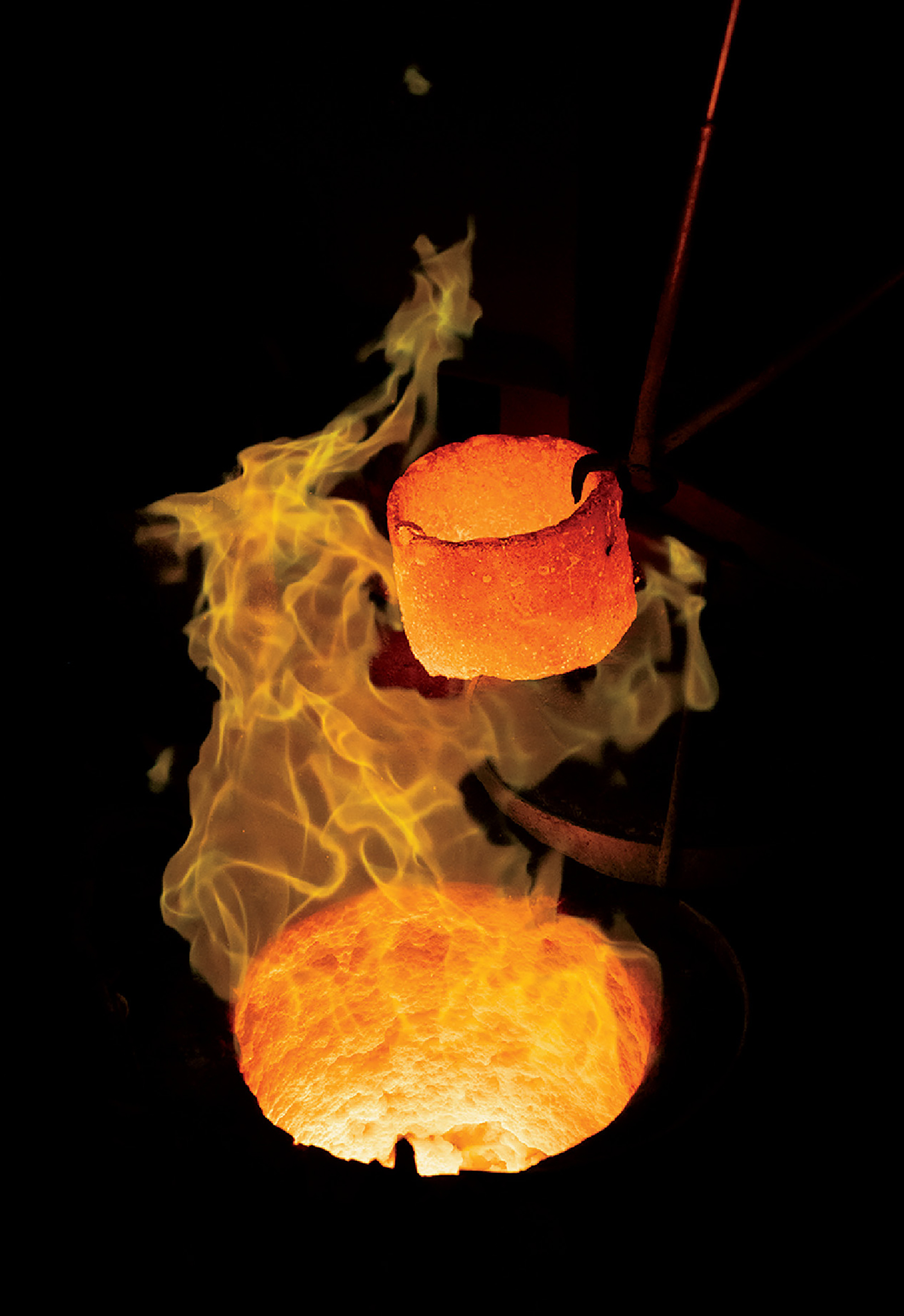 A heated crucible with glowing molten metal is lifted above a furnace, surrounded by bright orange flames and intense light.