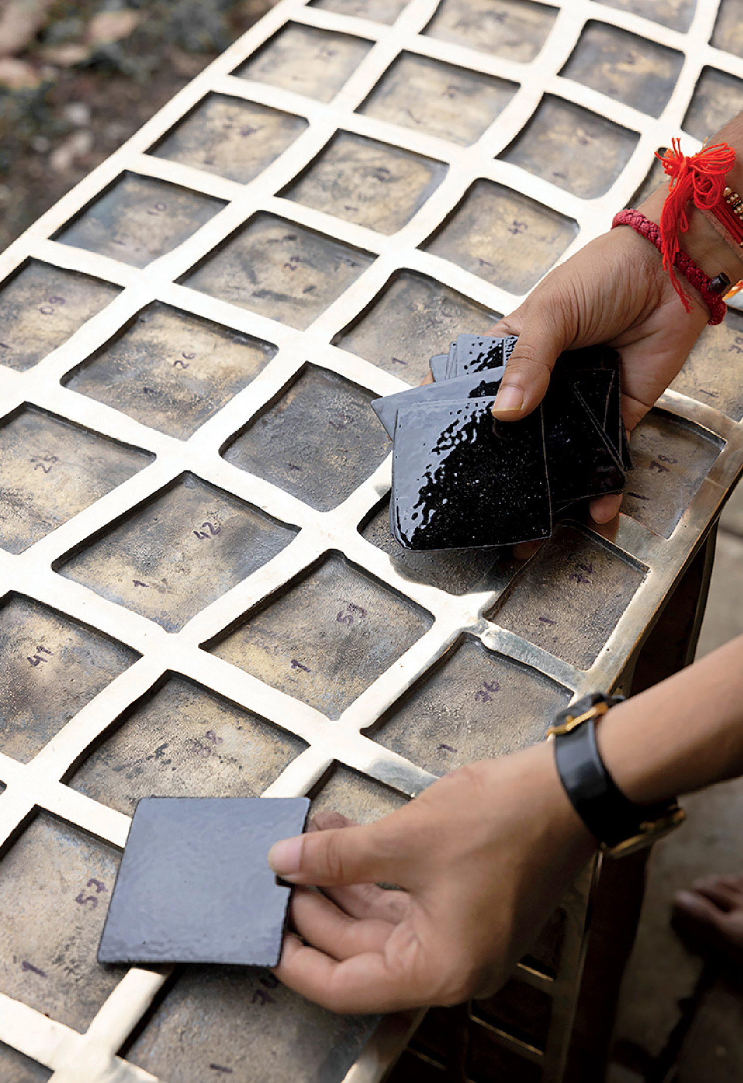 A person arranges glossy black tiles onto a metal grid marked with numbers, with hands protected by jewelry and a watch.