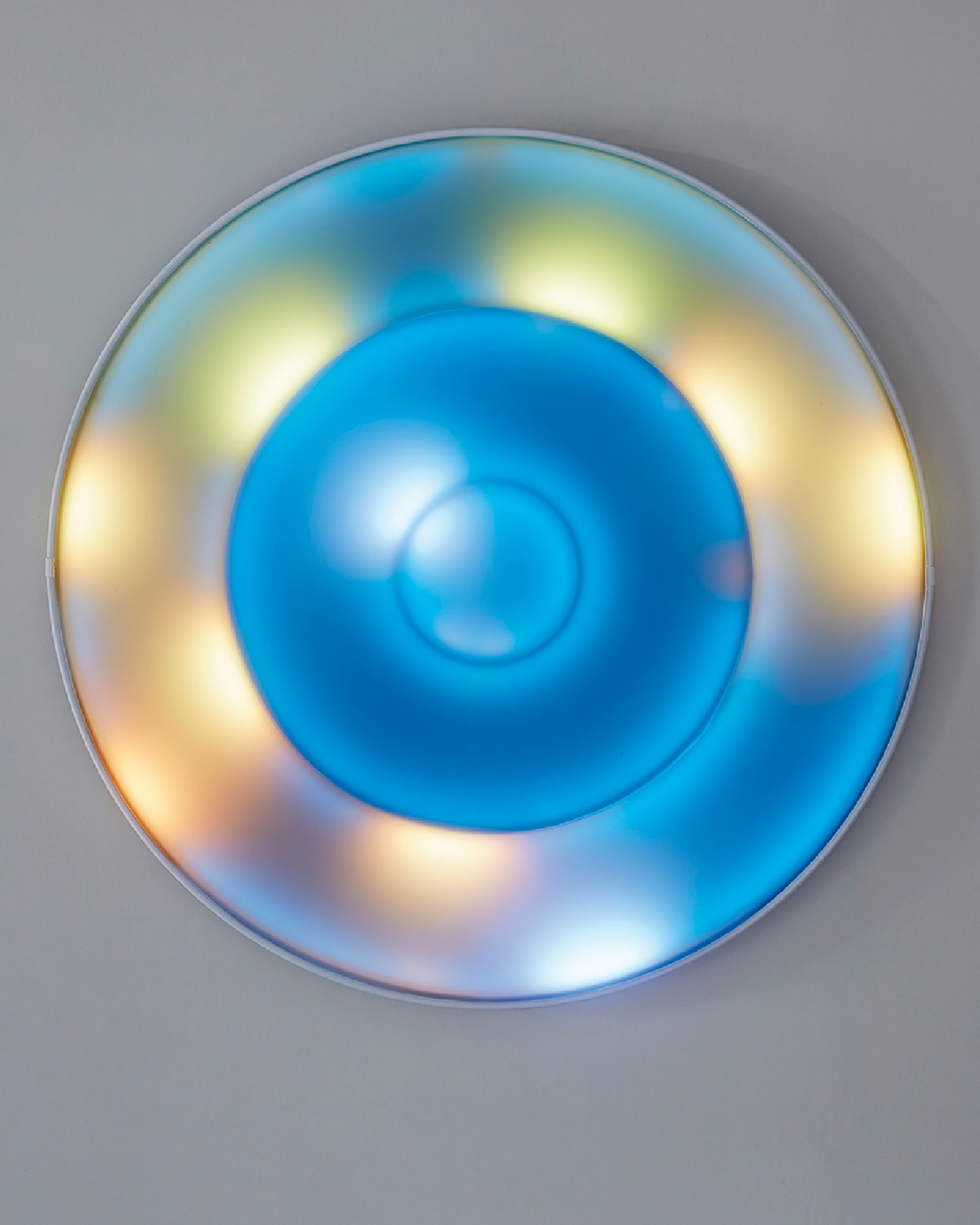 A round, blue-tinted wall fixture with concentric circles and soft, multicolored lights glowing around the edges, mounted on a gray wall.