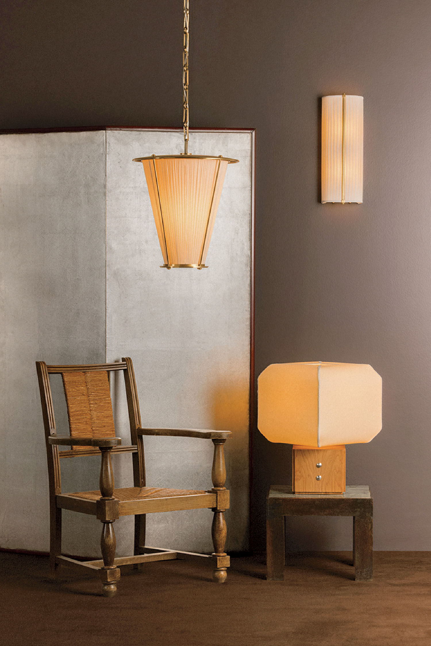 A wooden chair, a small table lamp, a hanging pendant light, and a wall sconce are arranged against a brown wall and a textured divider.