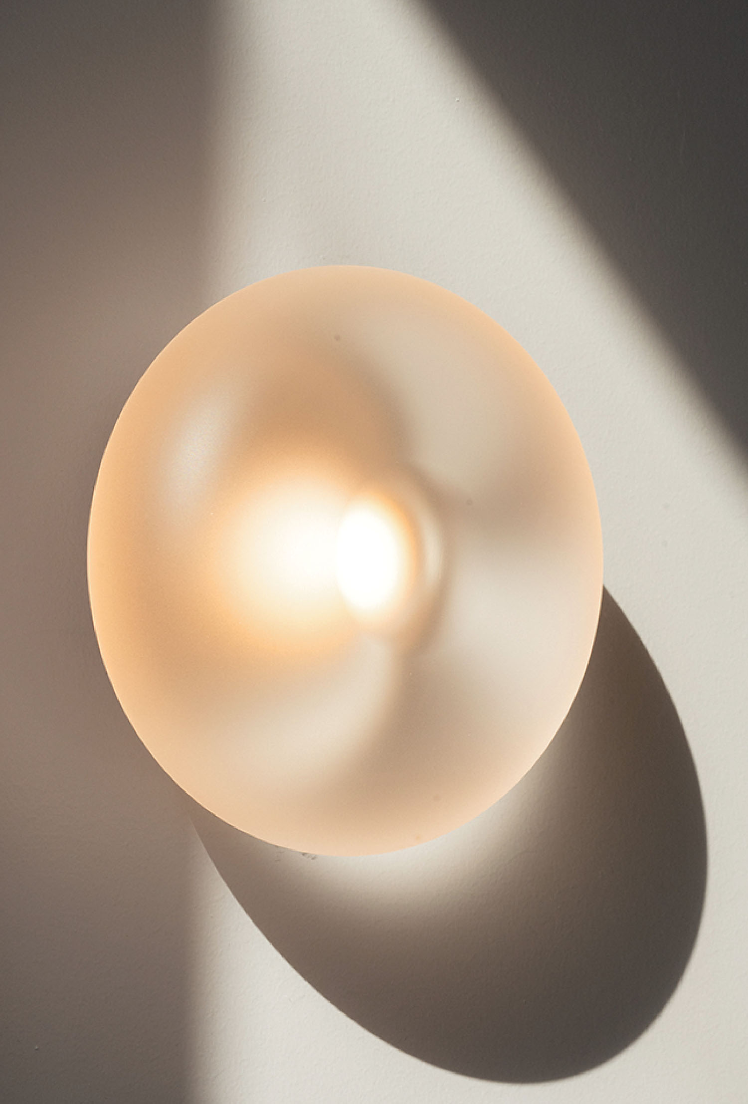 A round frosted glass light fixture mounted on a white wall, casting soft light and a distinct shadow to the right.