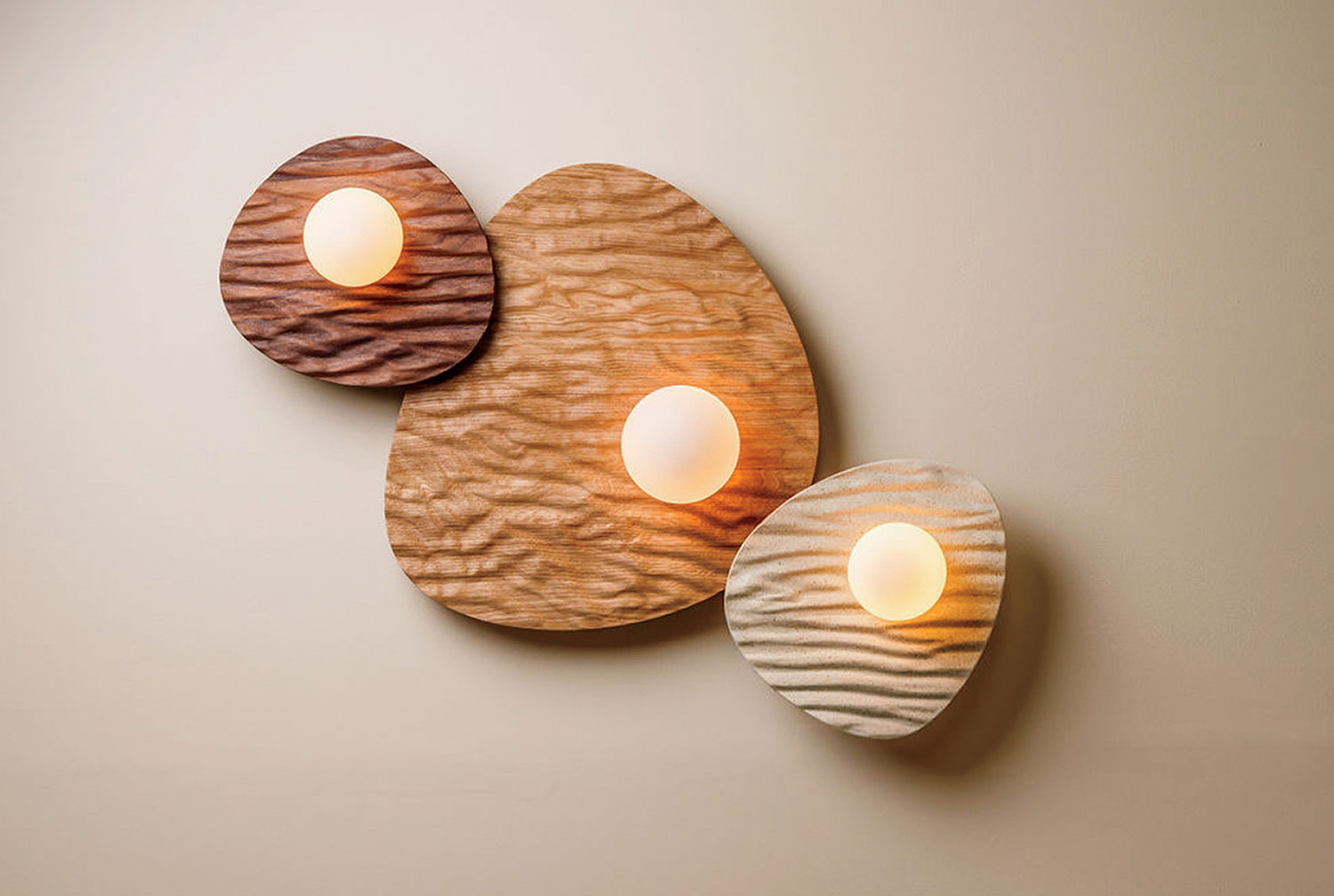 Three round wall lights are mounted on textured, organic-shaped wooden panels of varying sizes and colors against a plain wall.