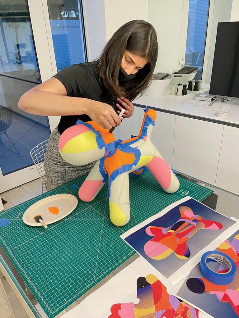 A woman applies colorful adhesive shapes to a dog-shaped sculpture on a cutting mat, with design plans and colored tape nearby on the table.