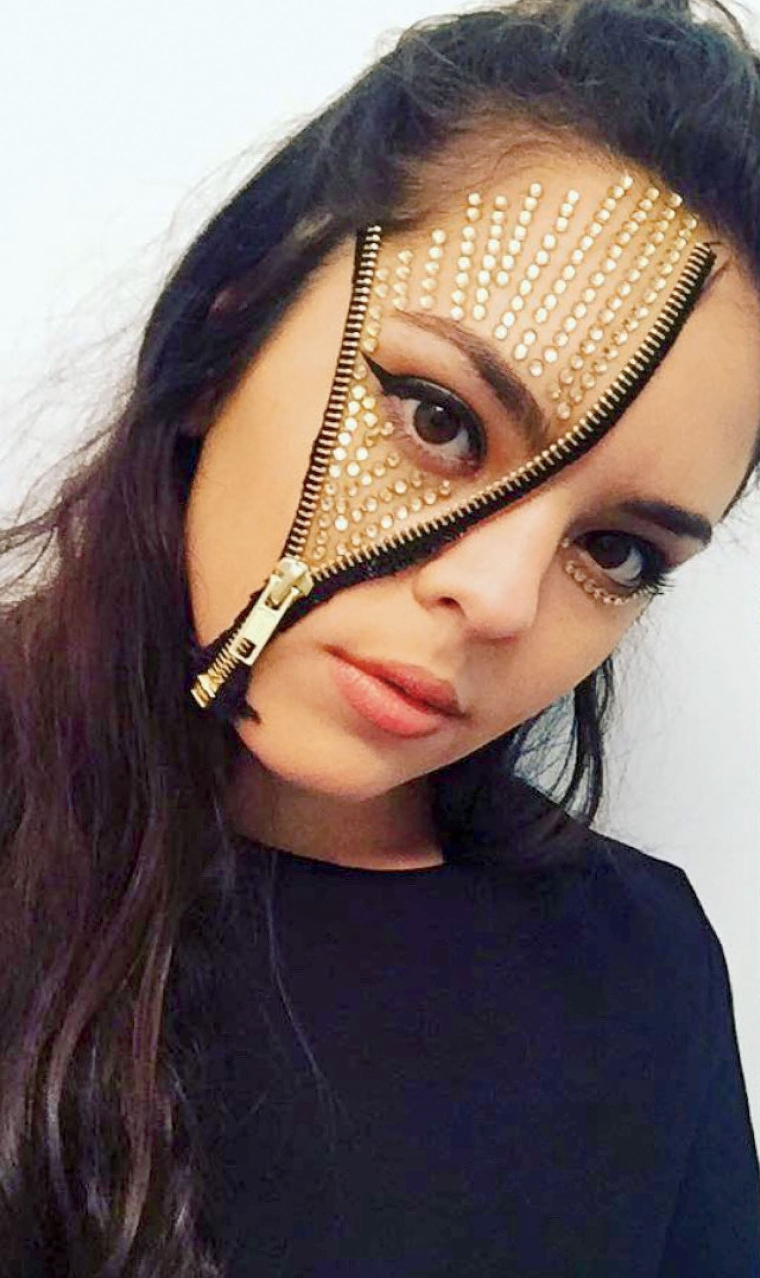 Woman with long dark hair wears makeup featuring a gold zipper over her face, revealing gold and rhinestone detailing around her eye and cheek.