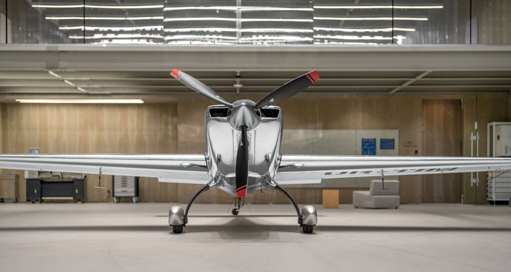A shiny, silver single-engine propeller aircraft is parked indoors in a modern hangar, viewed from the front.