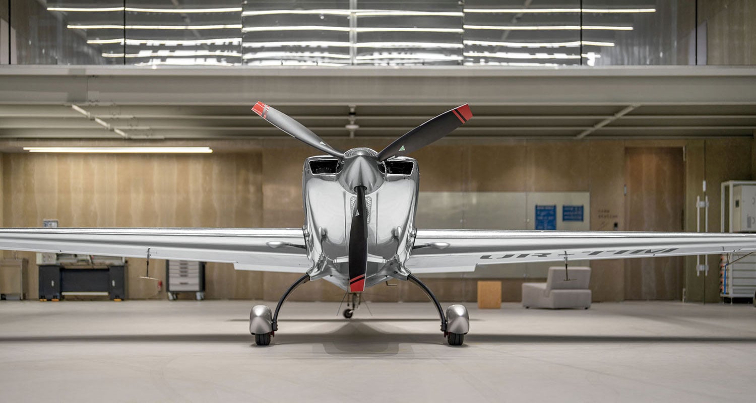 A shiny, silver single-engine propeller aircraft is parked indoors in a modern hangar, viewed from the front.