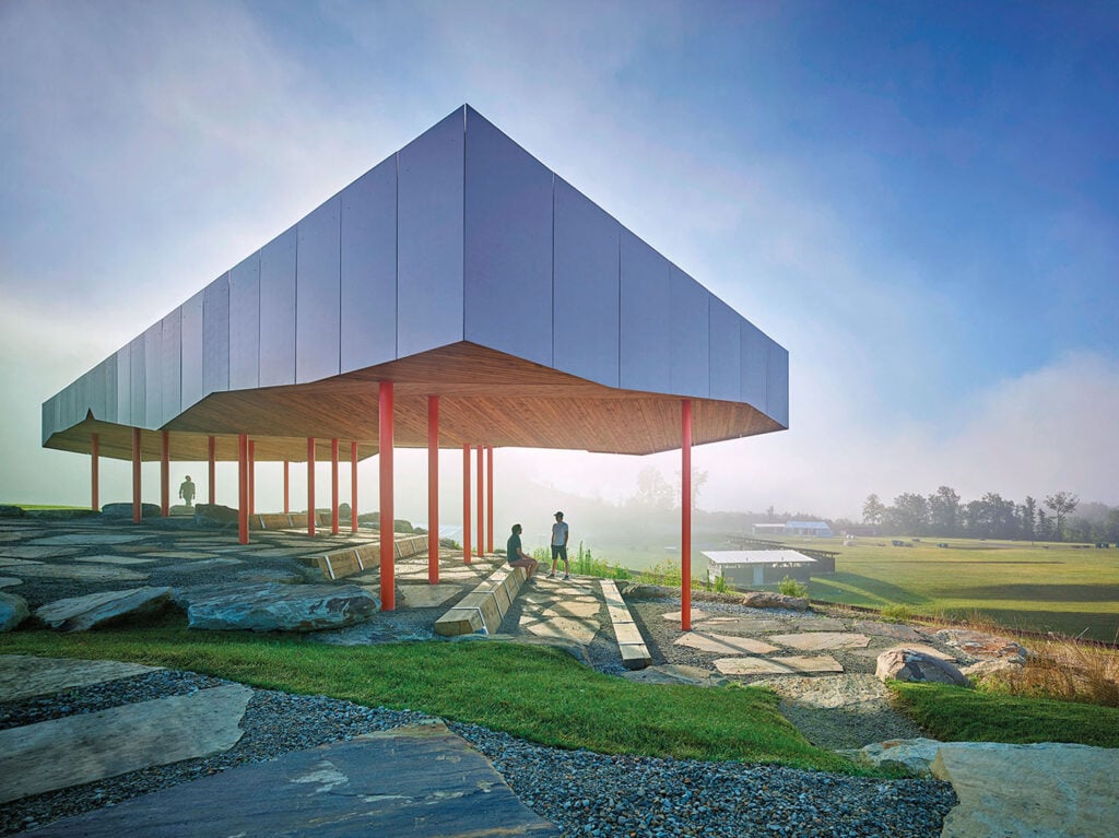 Modern pavilion with a geometric metal roof and red columns stands on a rocky landscape, with people sitting and walking nearby in a misty, open outdoor setting.