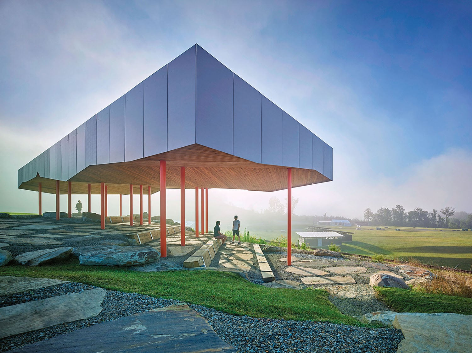 Modern pavilion with a geometric metal roof and red columns stands on a rocky landscape, with people sitting and walking nearby in a misty, open outdoor setting.