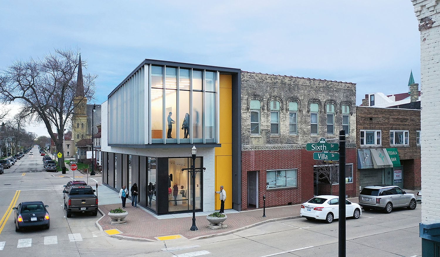 Modern two-story building with glass facade on a street corner, adjacent to older brick buildings, with people walking and cars parked nearby.