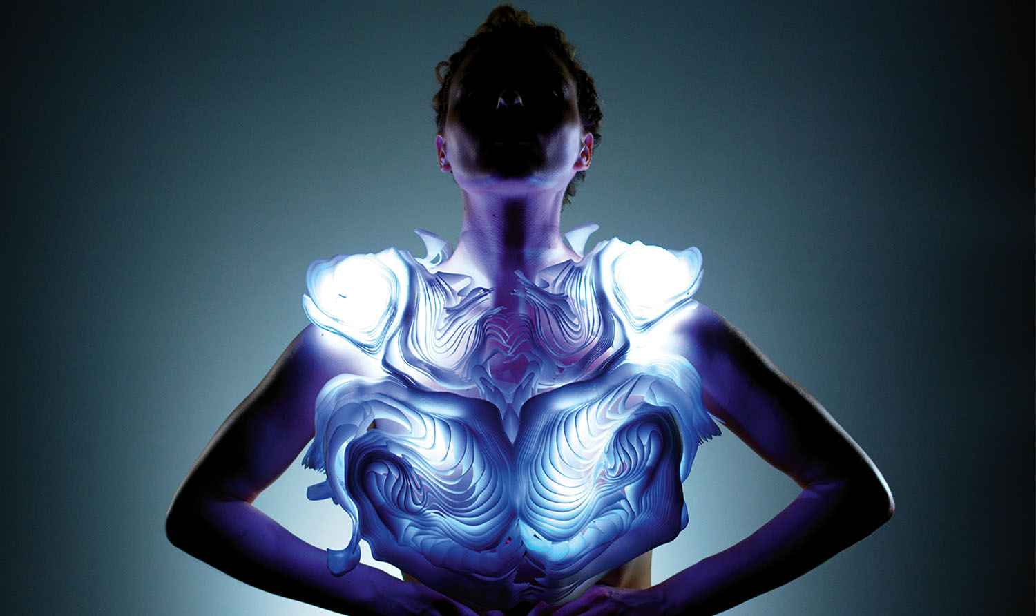 A person stands with hands on hips, wearing a futuristic, glowing sculptural garment that emits blue and white light, against a dark gradient background.
