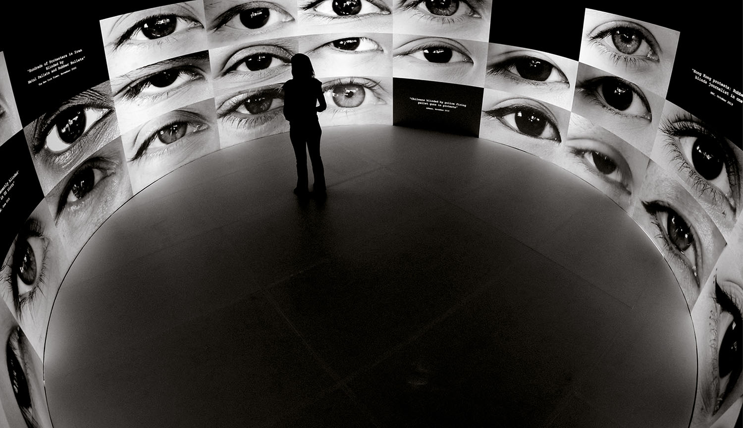A person stands in the center of a circular room surrounded by large screens displaying close-up images of human eyes.