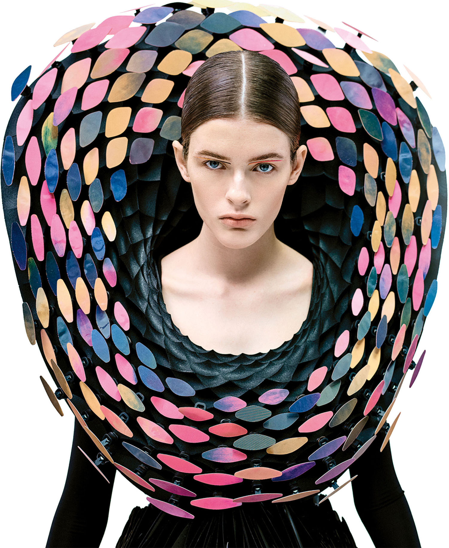 A person with slicked-back hair wears a dramatic black outfit featuring a large, circular collar decorated with colorful, oval-shaped pieces.