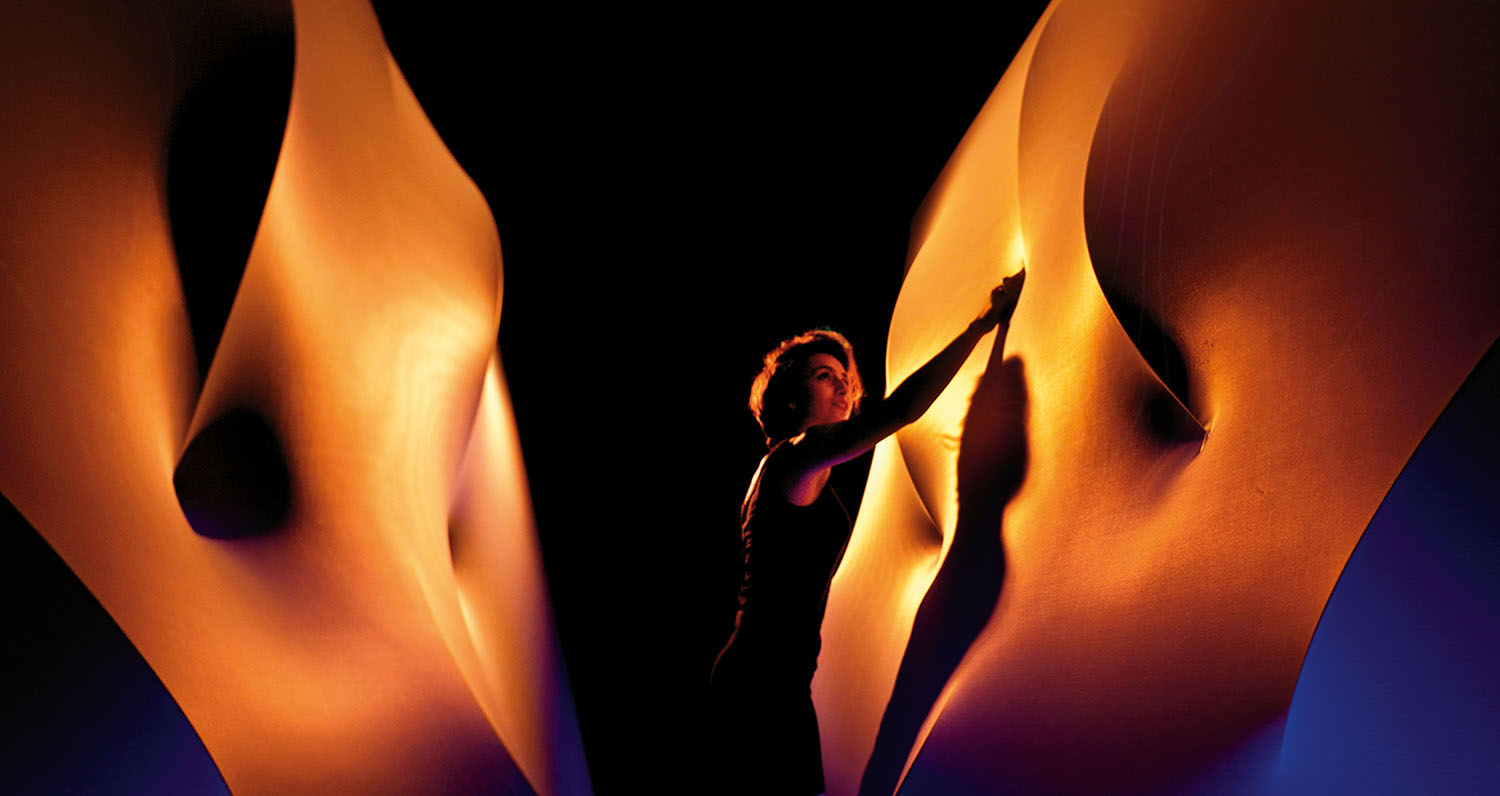 A person stands between two large, curved, orange-lit fabric structures, pressing their hands against one surface in a dark setting.