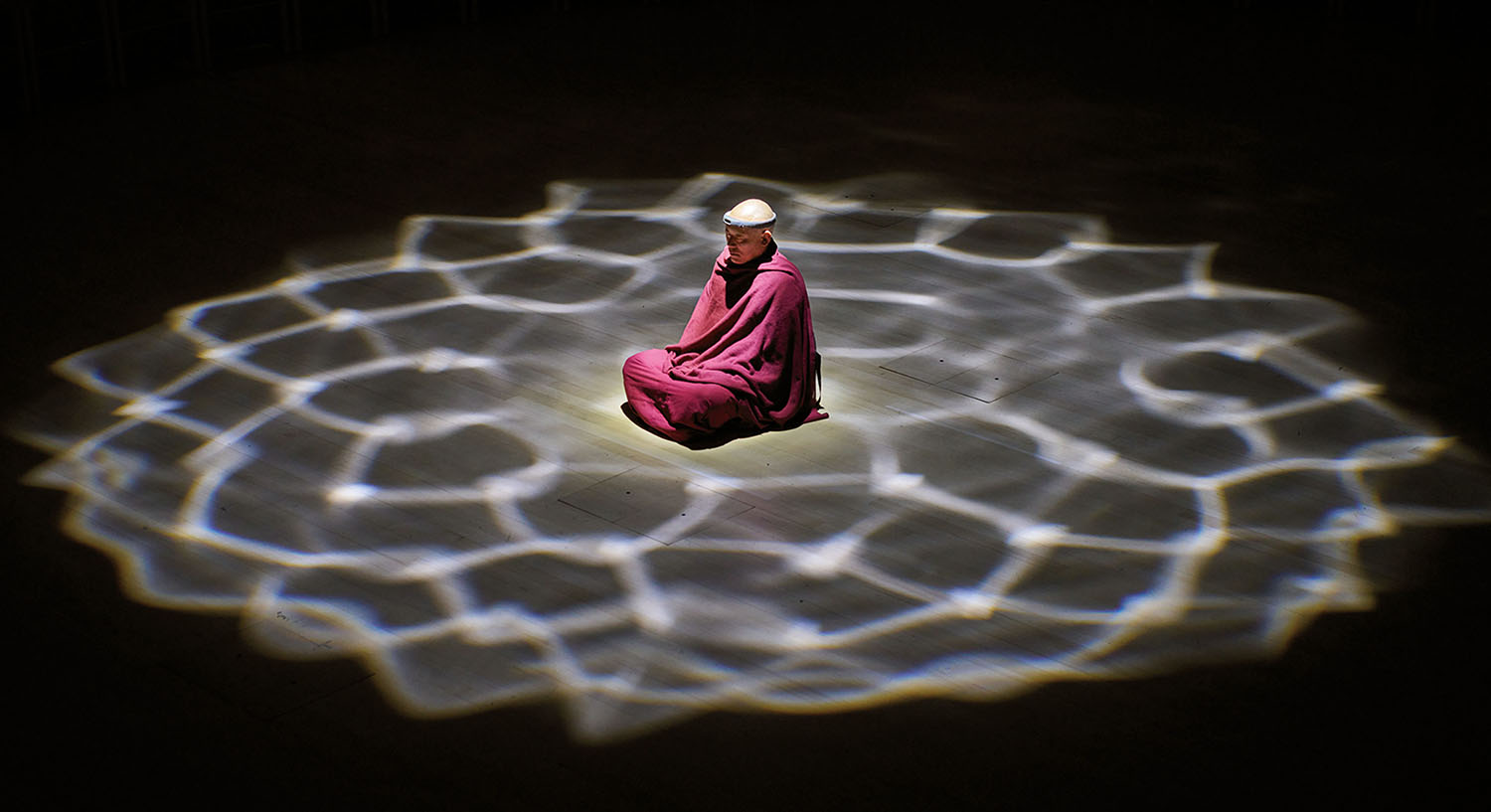 A person in a maroon robe sits cross-legged on the floor, illuminated by a patterned circular spotlight against a dark background.