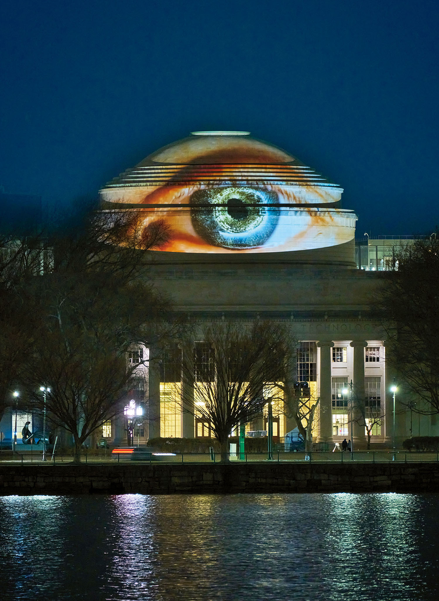 A large, realistic image of a human eye is projected onto the domed roof of a neoclassical building at night, with trees and water in the foreground.