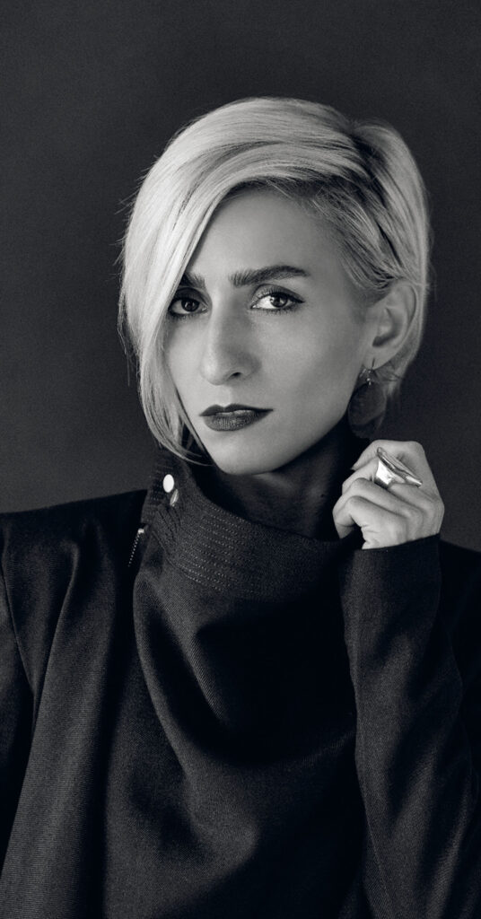 Black and white portrait of a person with short, light hair wearing a dark high-collared top, looking at the camera with a serious expression and holding their collar.