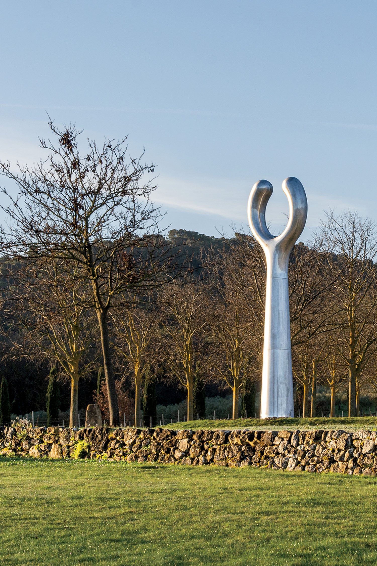 A tall, polished metal sculpture with two upward-curving arms stands on a stone platform amid bare trees and grass, with hills in the background.