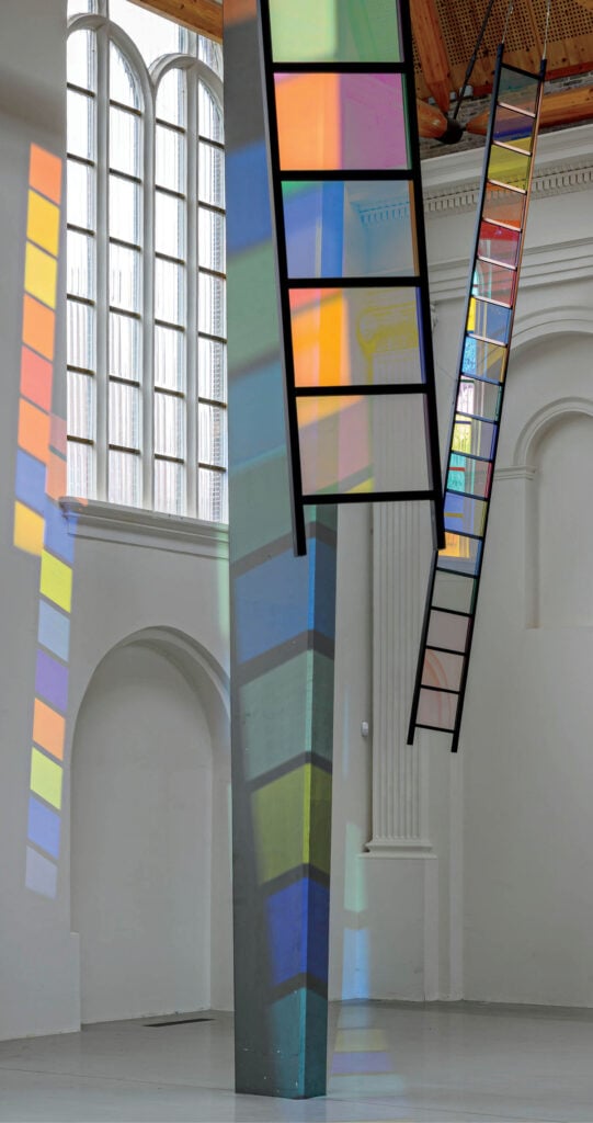 Tall, colorful glass panels hang in a white gallery, casting multicolored shadows and reflections onto the floor, walls, and columns.