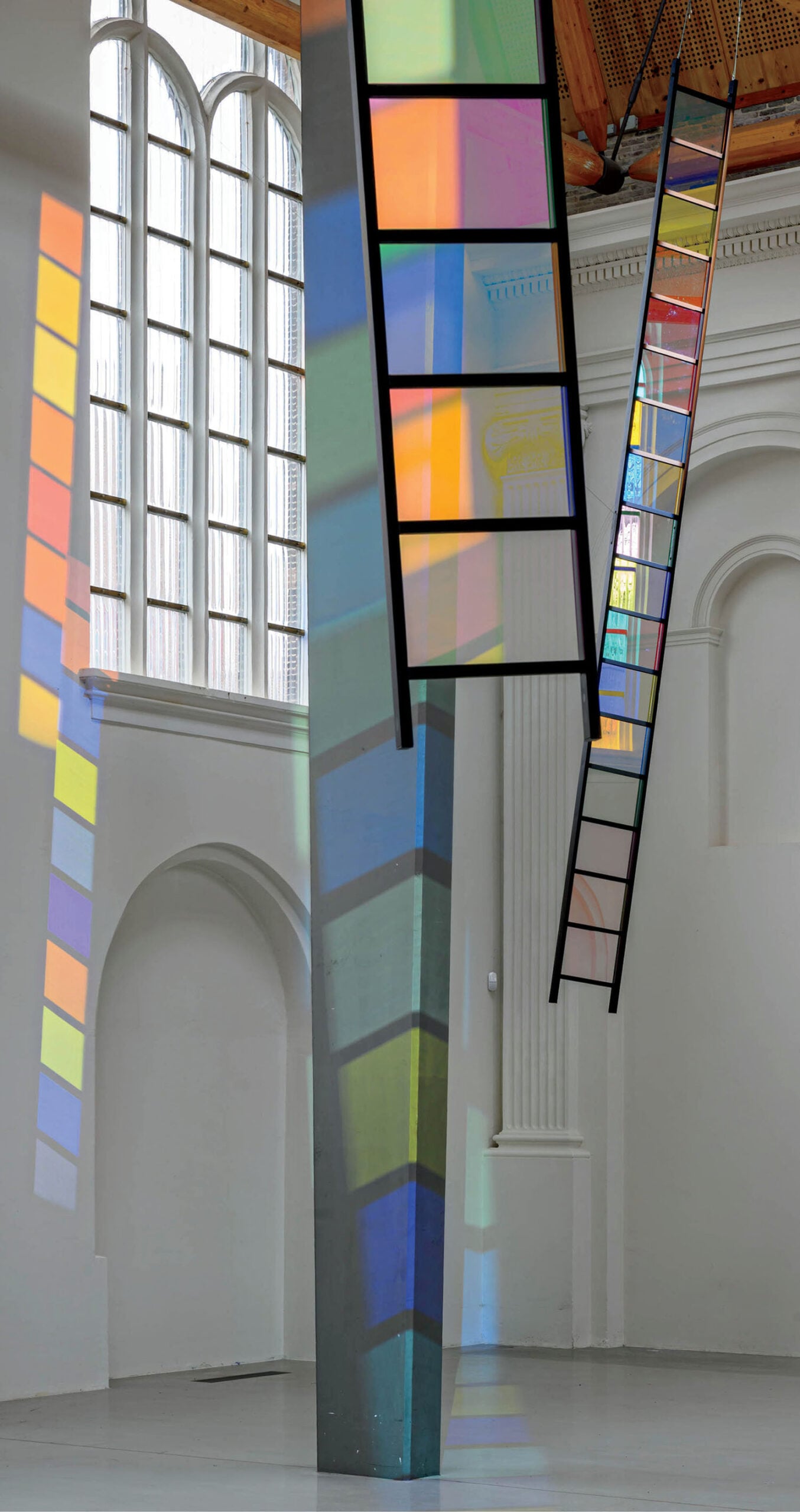 Tall, colorful glass panels hang in a white gallery, casting multicolored shadows and reflections onto the floor, walls, and columns.