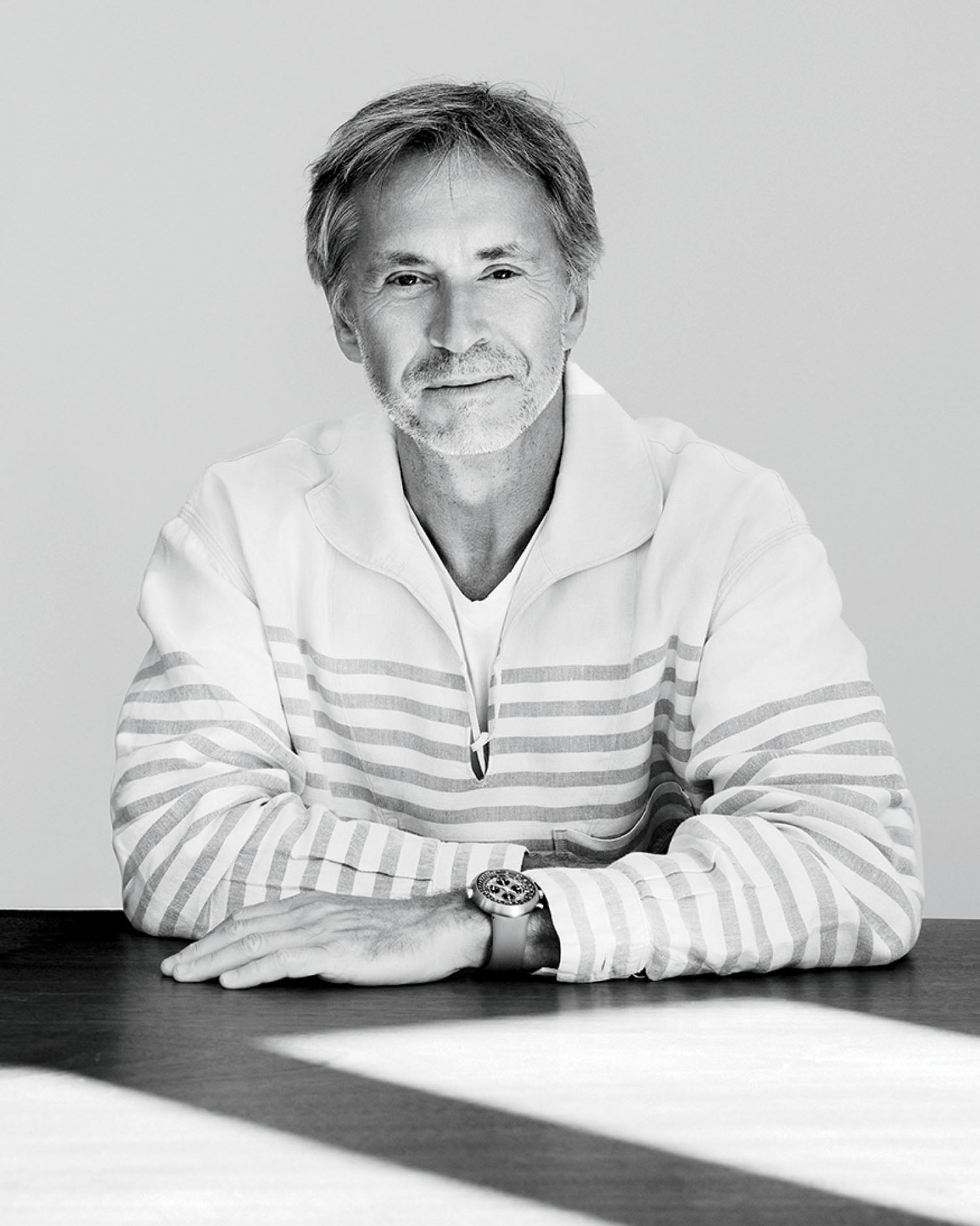 A man with gray hair and a beard sits at a table, wearing a light-colored, striped pullover and a wristwatch, looking at the camera with a slight smile.