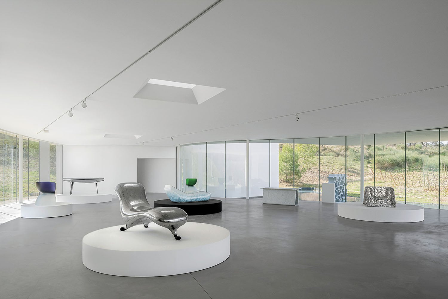Modern gallery space with large windows displaying various contemporary furniture pieces on white circular platforms.