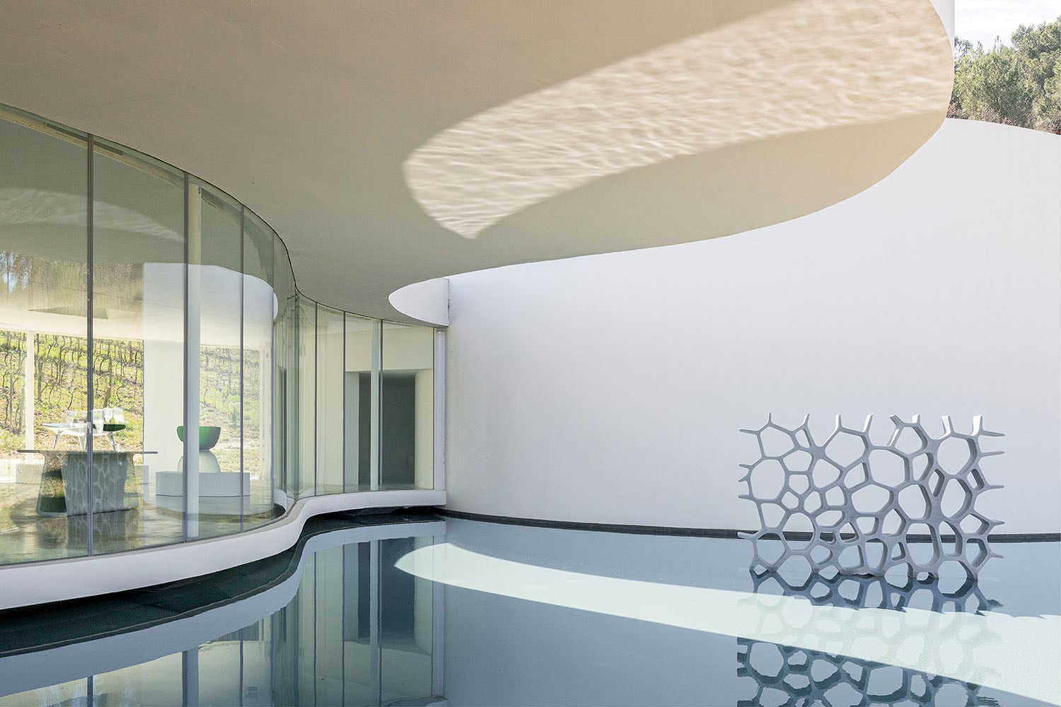 Modern architectural space with curved glass walls, white ceiling, reflecting pool, and a geometric sculpture near the water.