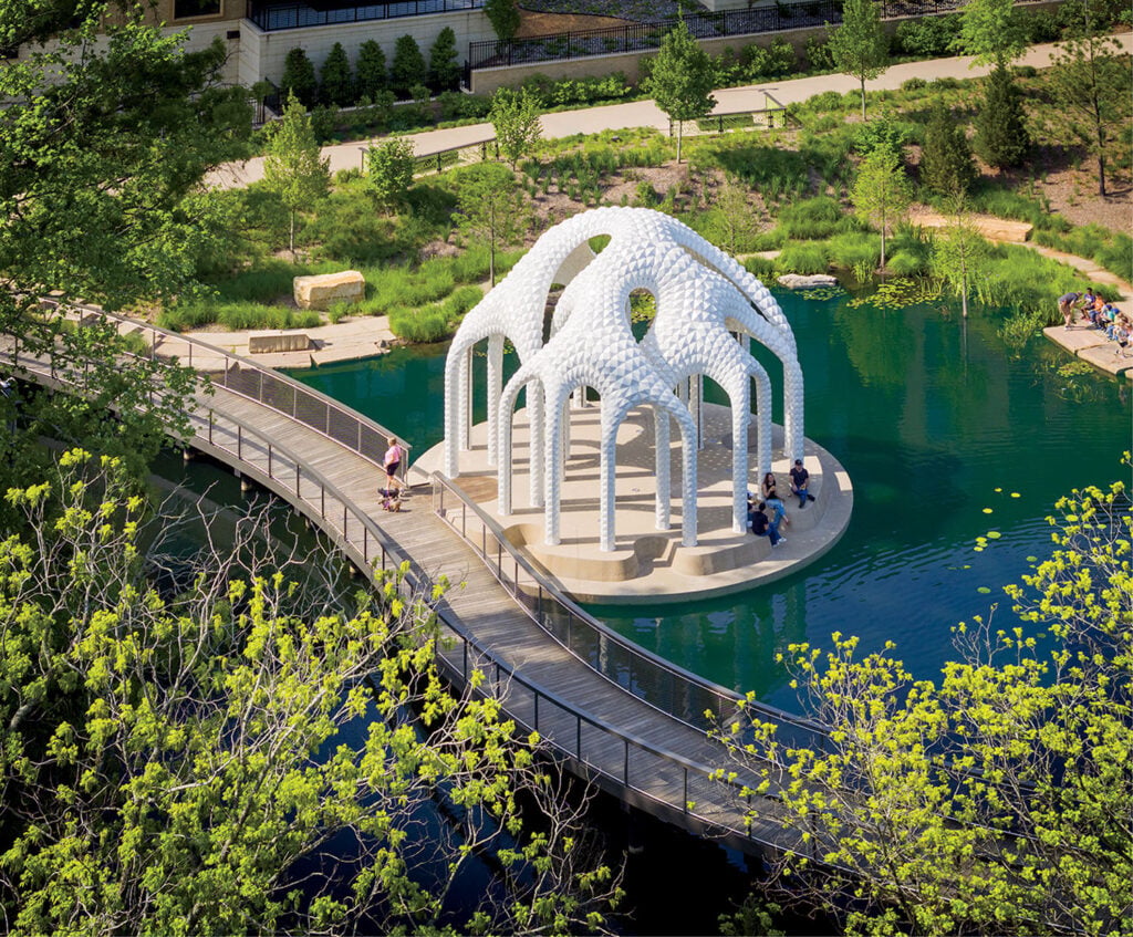 A footbridge crosses a pond to a white geometric pavilion surrounded by greenery, with people walking and sitting nearby.