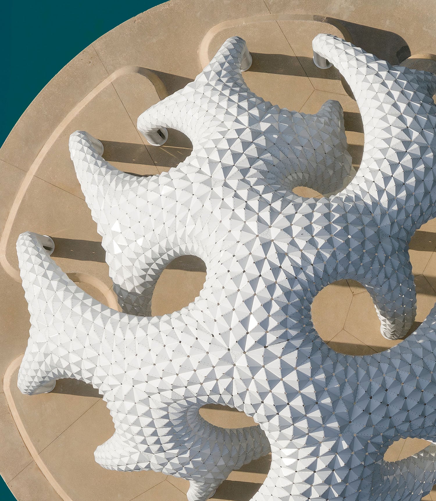 White geometric, spiky sculpture with circular cutouts viewed from above, set against a beige, circular concrete surface and a teal background.
