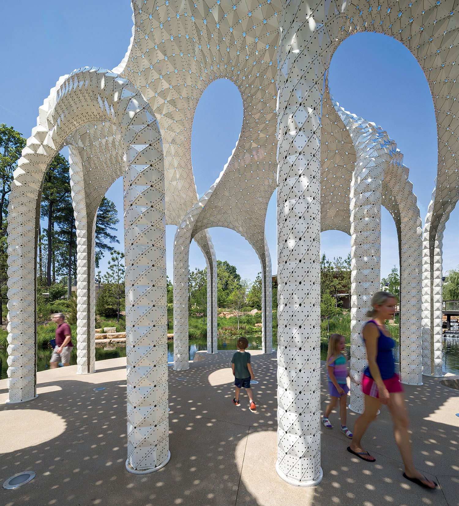 People walk under a large outdoor pavilion with tall, perforated white columns and arches casting patterned shadows on the ground. Trees and water are visible in the background.