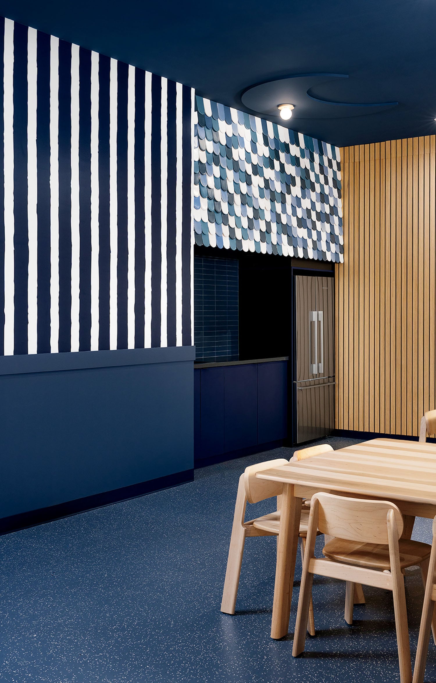 A modern room with navy and white vertical stripes, a wall with blue and white scale-like tiles, wooden slats, a wooden table, and chairs on a speckled blue floor.