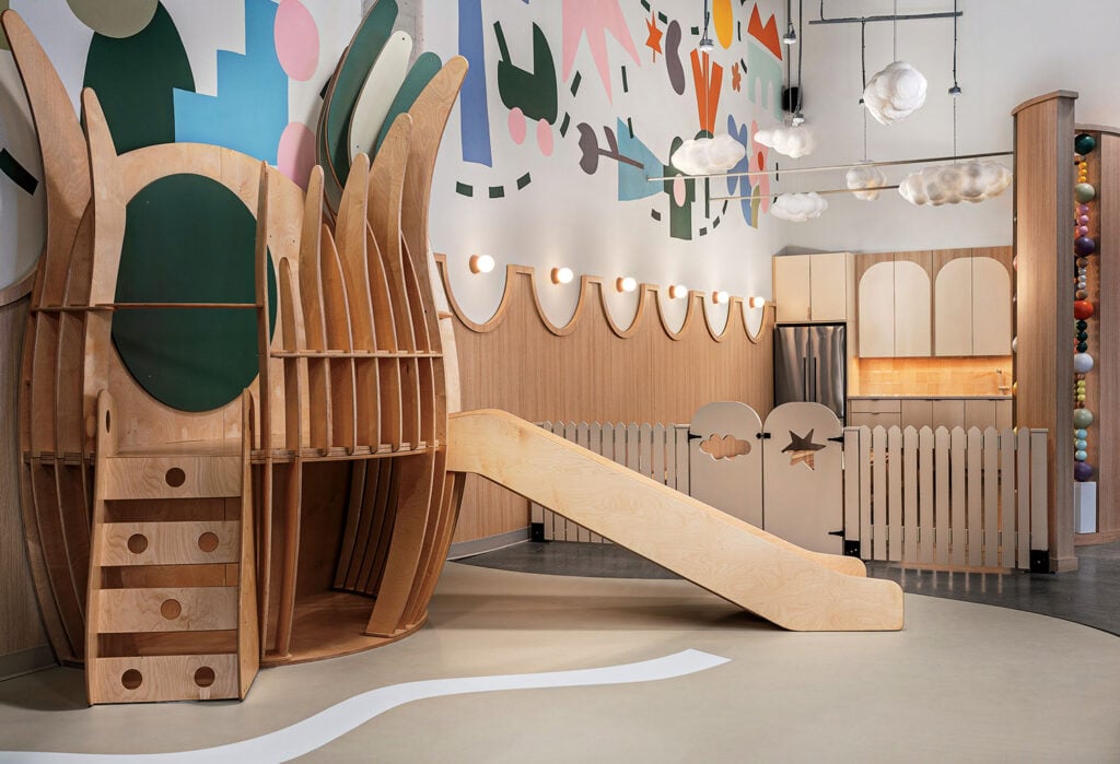 Modern indoor playroom with wooden climbing structure, slide, play kitchen area, ball pit, and colorful abstract wall art.