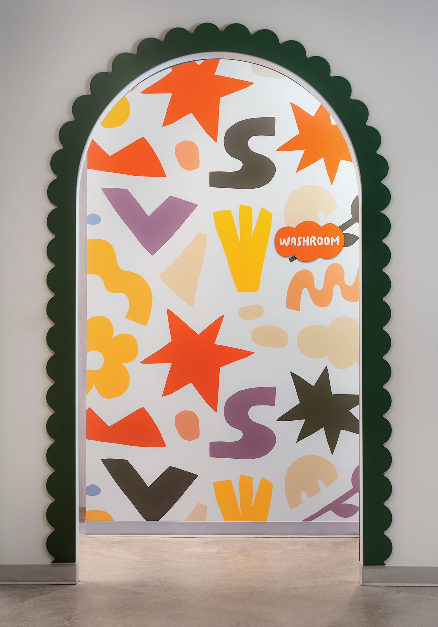 An arched doorway with a scalloped green frame opens to a colorful wall featuring abstract shapes, stars, and the word "WASHROOM" in orange text.
