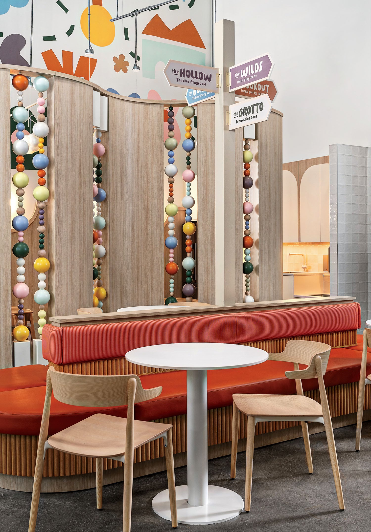 Colorful modern café interior with wooden chairs, a white round table, red upholstered bench seating, beaded room dividers, decorative signs, and playful wall art.