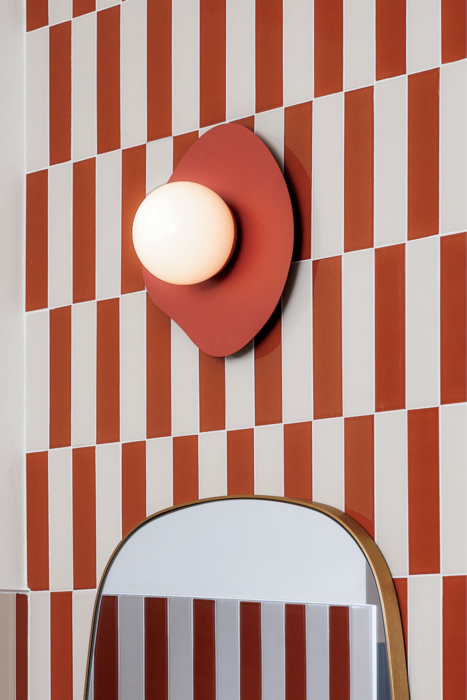 A modern wall lamp with a round white bulb and red base is mounted on a wall with vertical red and white rectangular tiles above a mirror.