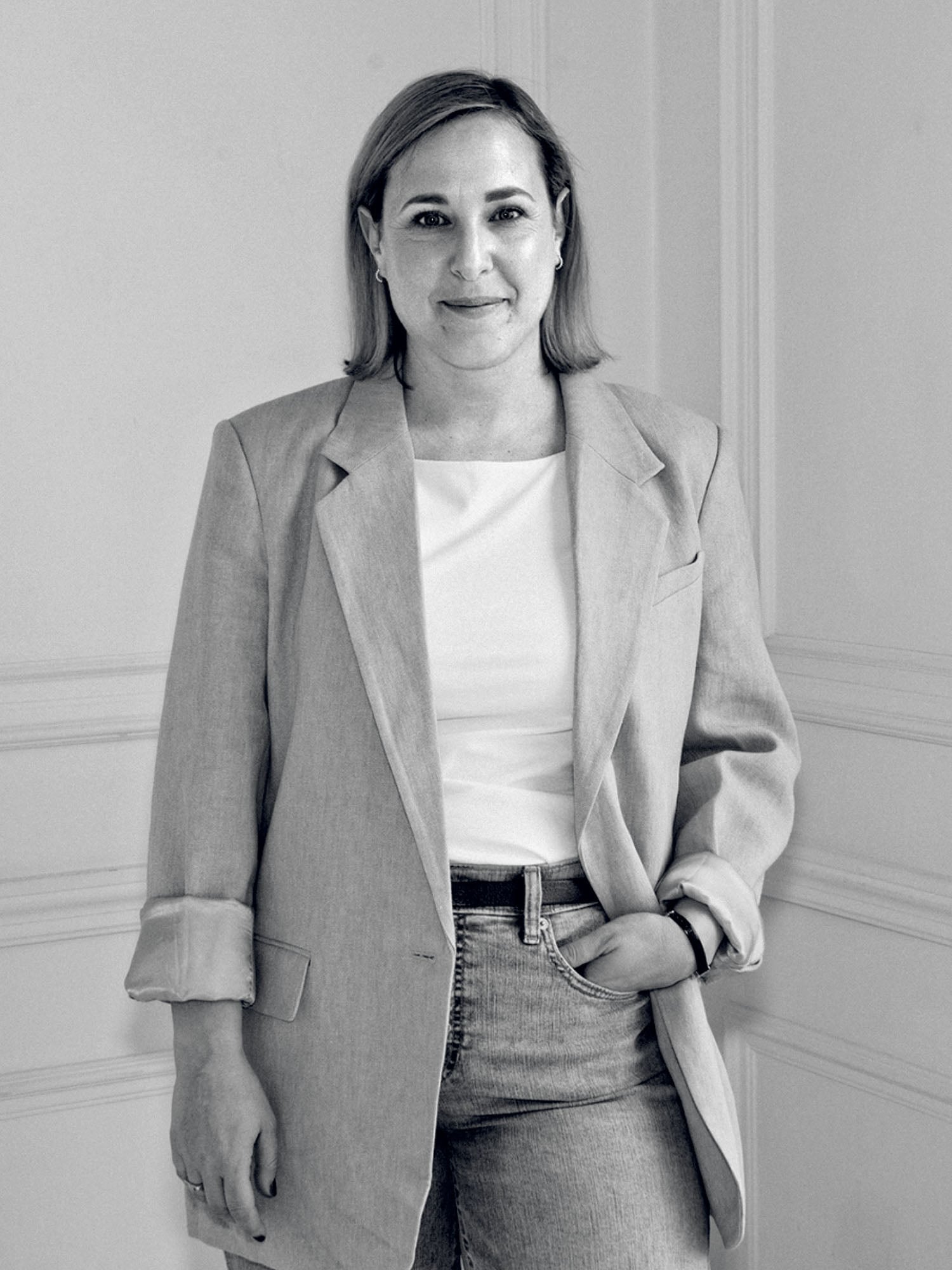 A woman stands indoors against a wall, wearing a light blazer, white top, and jeans, with one hand in her pocket and a neutral expression.
