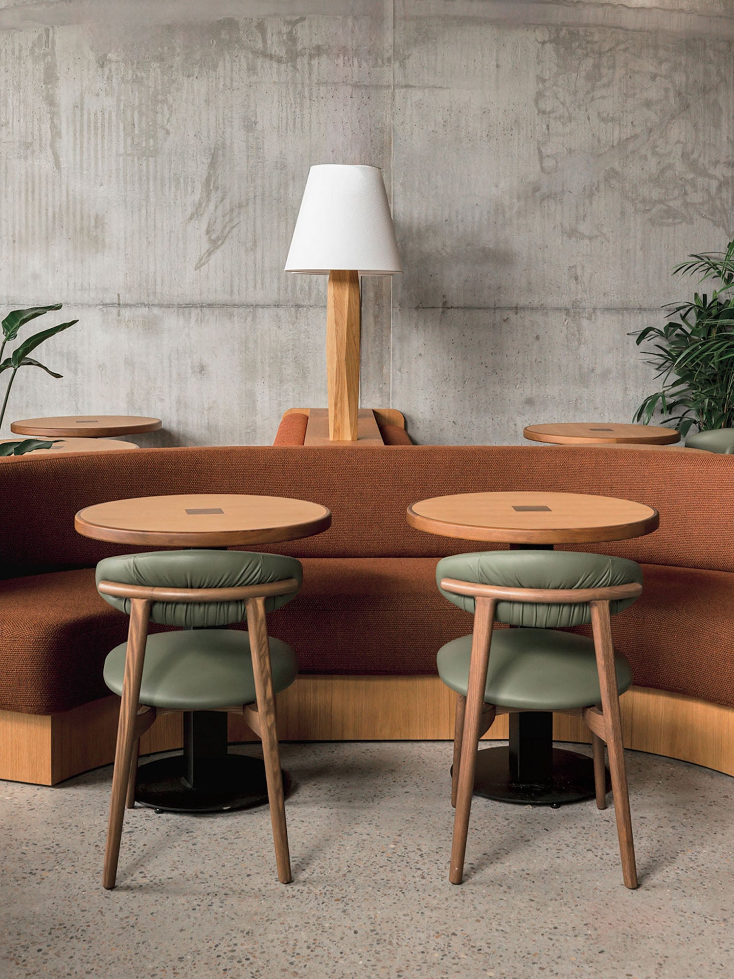 Two round wooden tables with green cushioned stools sit in front of a curved rust-colored sofa, with a wooden lamp and plants against a concrete wall.