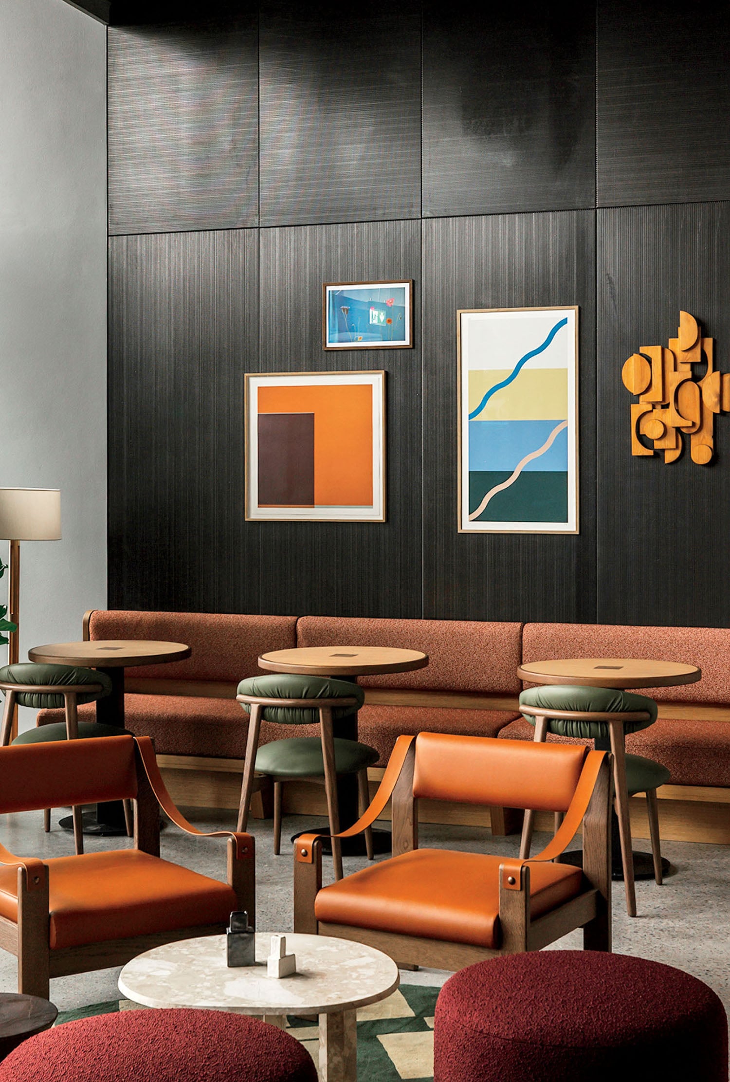 Modern lounge with brown leather chairs, round tables, a long upholstered bench, abstract wall art, and a dark wood panel backdrop.