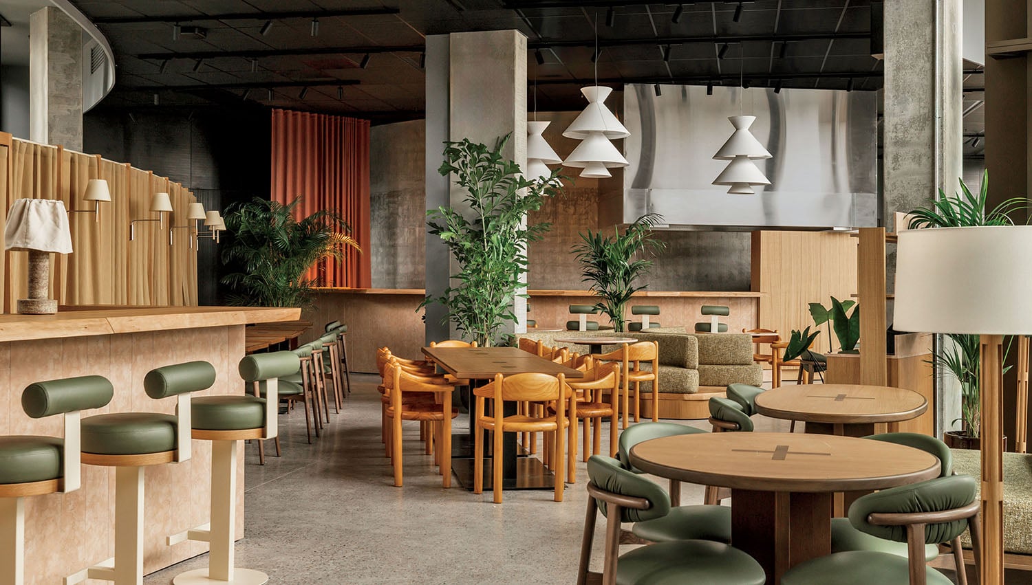 Modern restaurant interior with wooden tables, green and tan chairs, pendant lights, plants, and neutral-colored walls and floors.