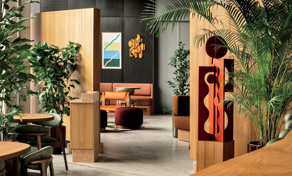 Modern lounge with wood partitions, green plants, abstract art on dark walls, and contemporary furniture in earthy tones arranged throughout the space.