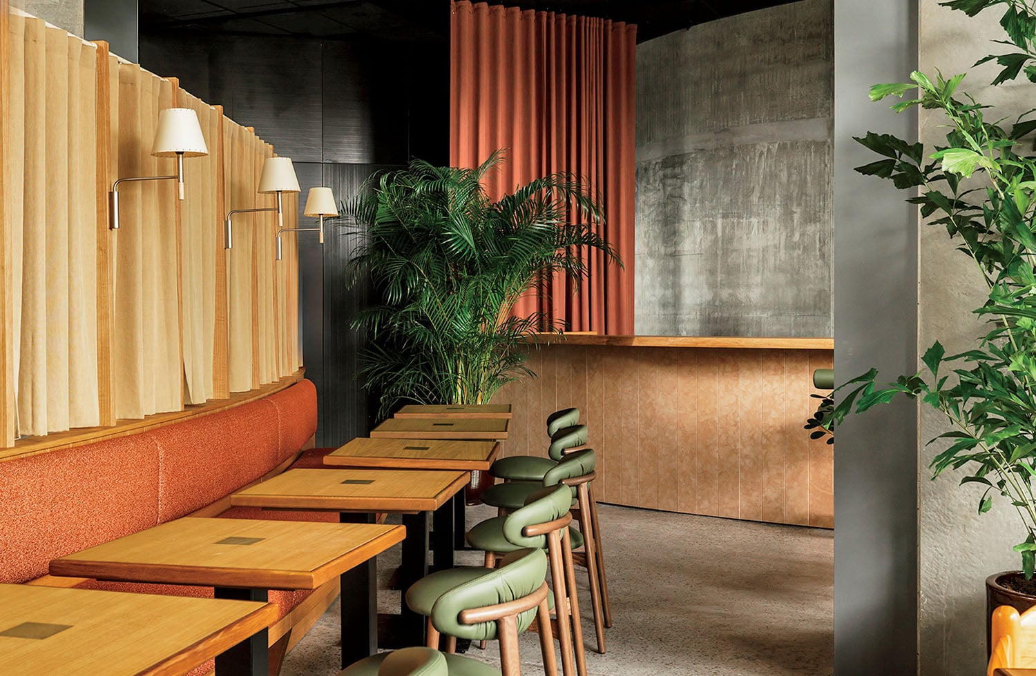 A modern cafe interior with wooden tables, green and brown chairs, fabric panels, potted plants, and a textured concrete wall.