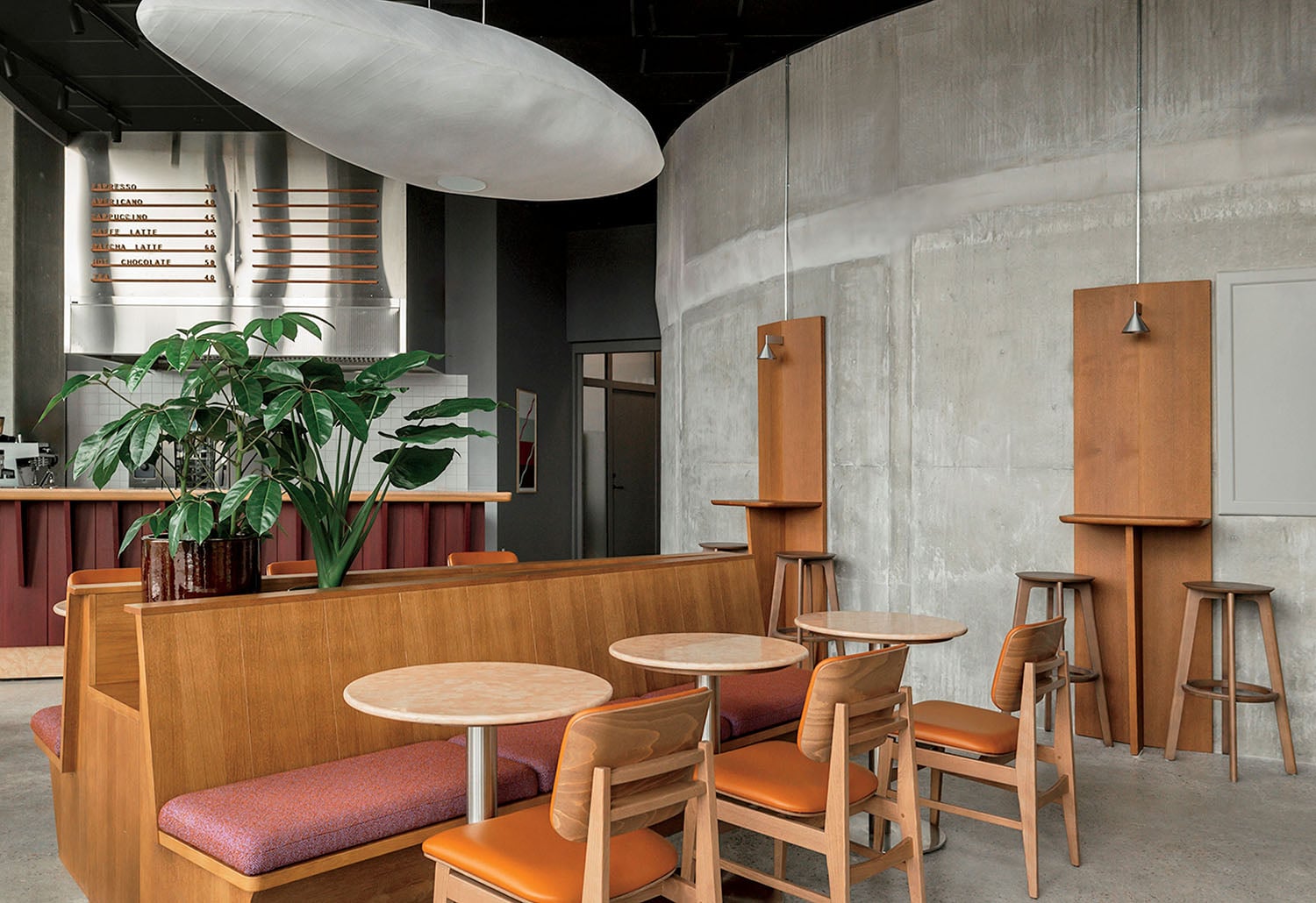 Modern café interior with wooden tables and chairs, a built-in planter, curved concrete wall, pendant lights, and a counter with a menu board in the background.