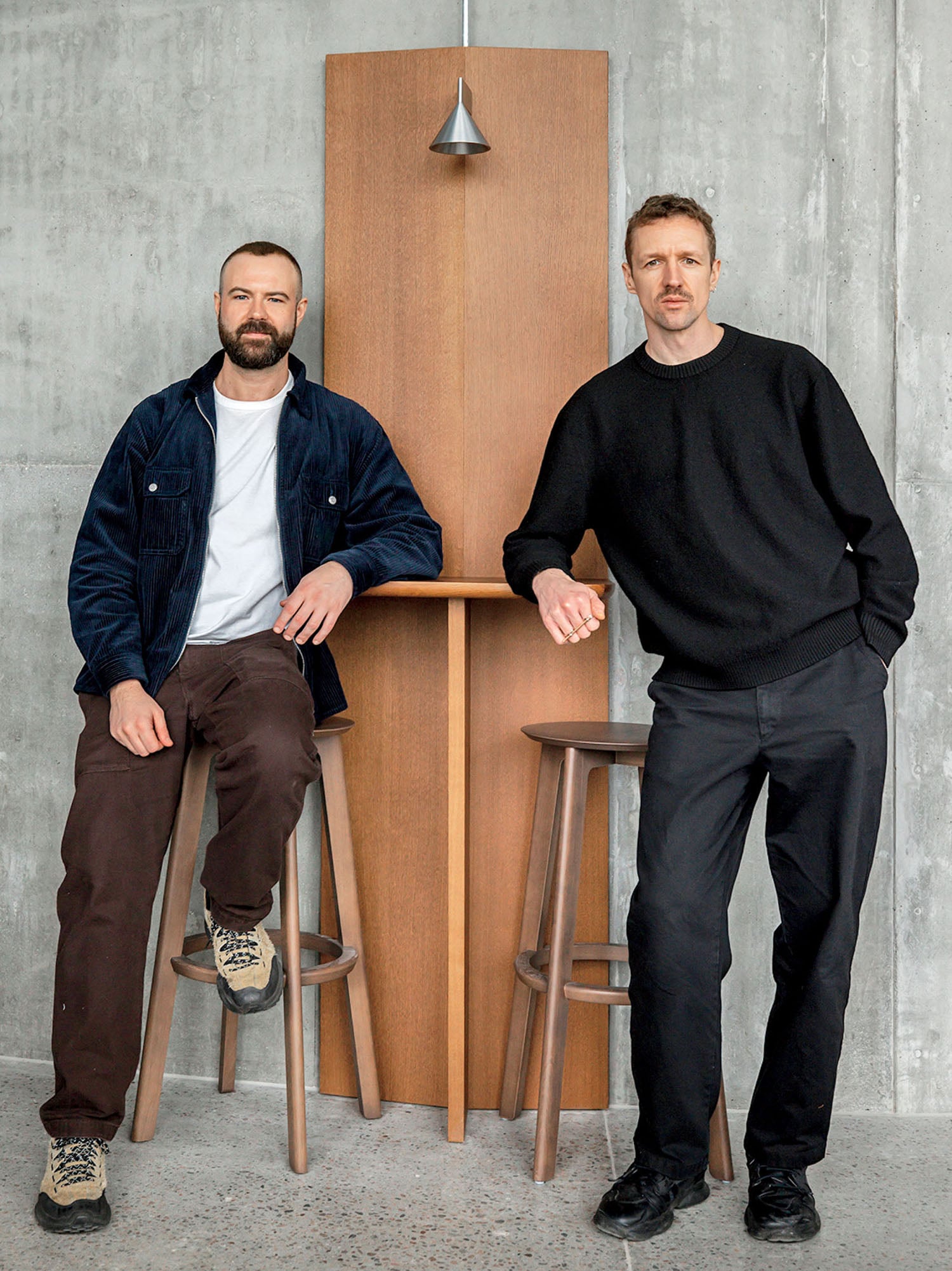 Two men pose in front of a concrete wall, standing and sitting by a tall wooden table with two stools and a hanging pendant light above.