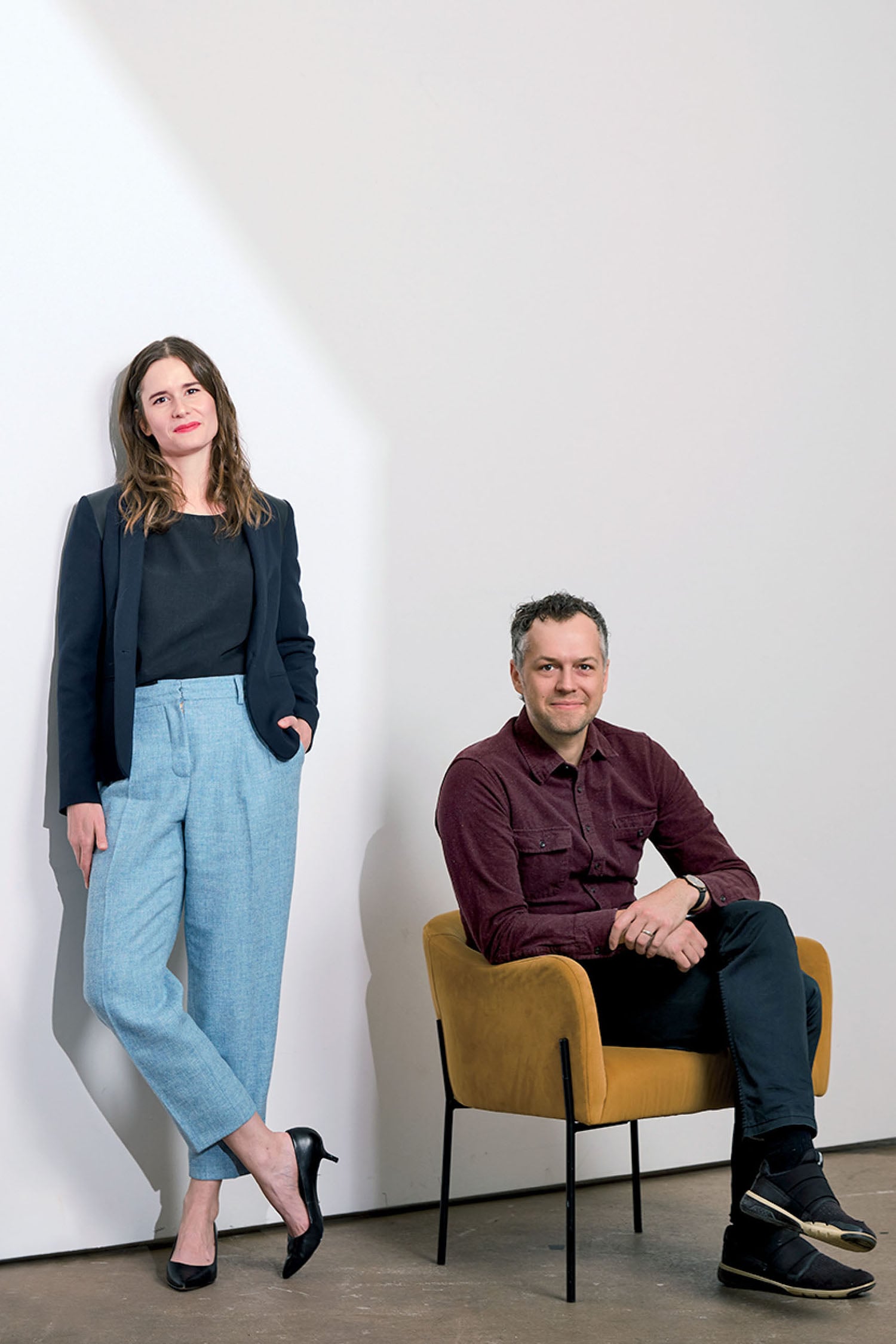 A woman stands with hands in her pockets next to a man seated in a yellow chair against a plain white wall.