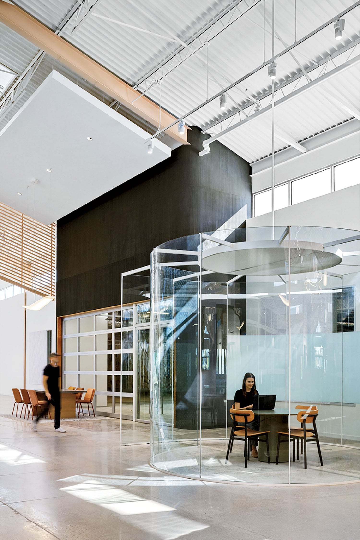 A modern office space with glass meeting rooms, high ceilings, and natural light; one person sits at a desk inside a glass enclosure while another walks past outside.
