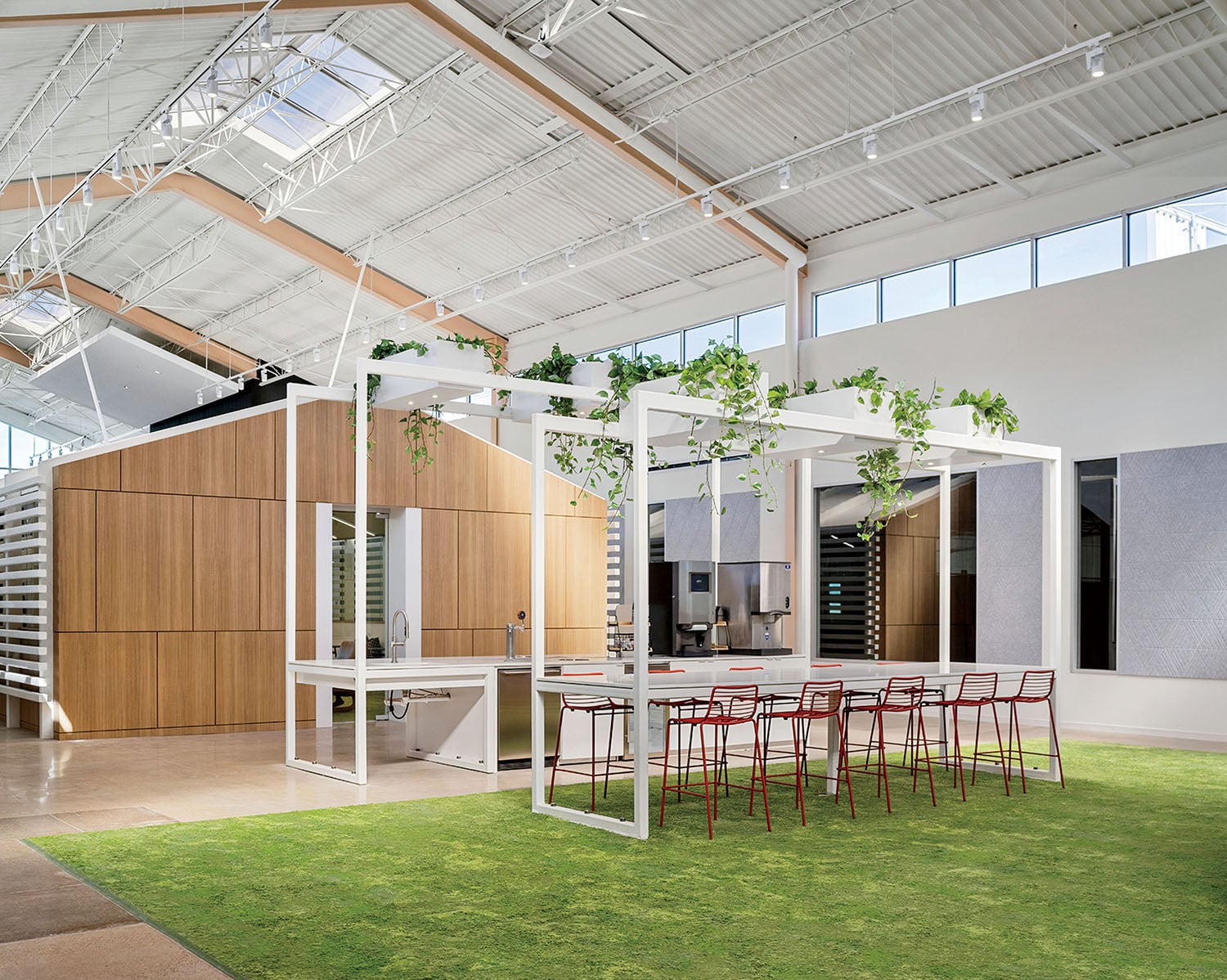 Modern office space with a communal table, red chairs, overhead white frame with hanging plants, and natural light from skylights.