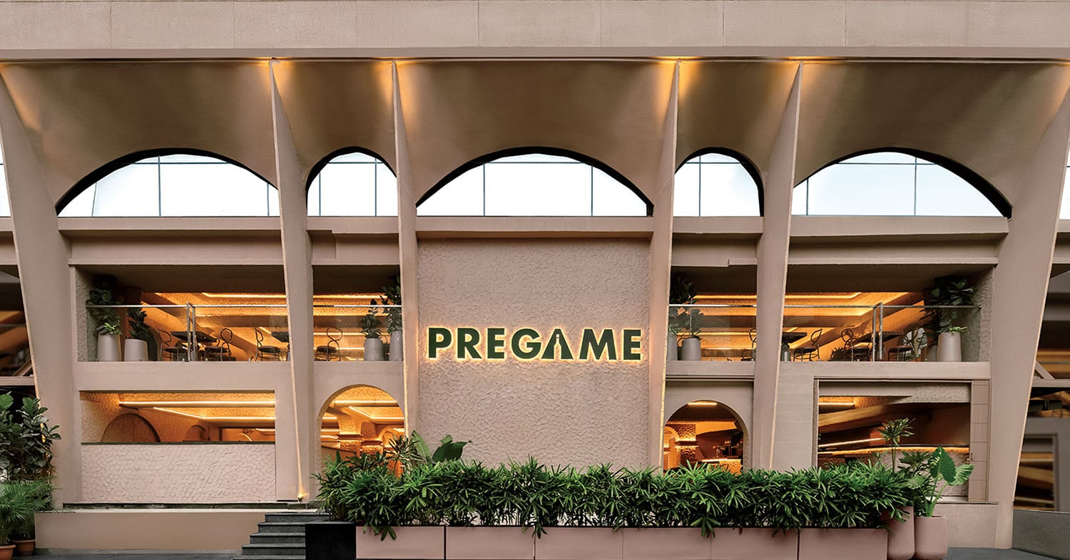 The exterior of a modern building with large arched windows, greenery at the entrance, and a sign reading "PREGAME" on the facade.
