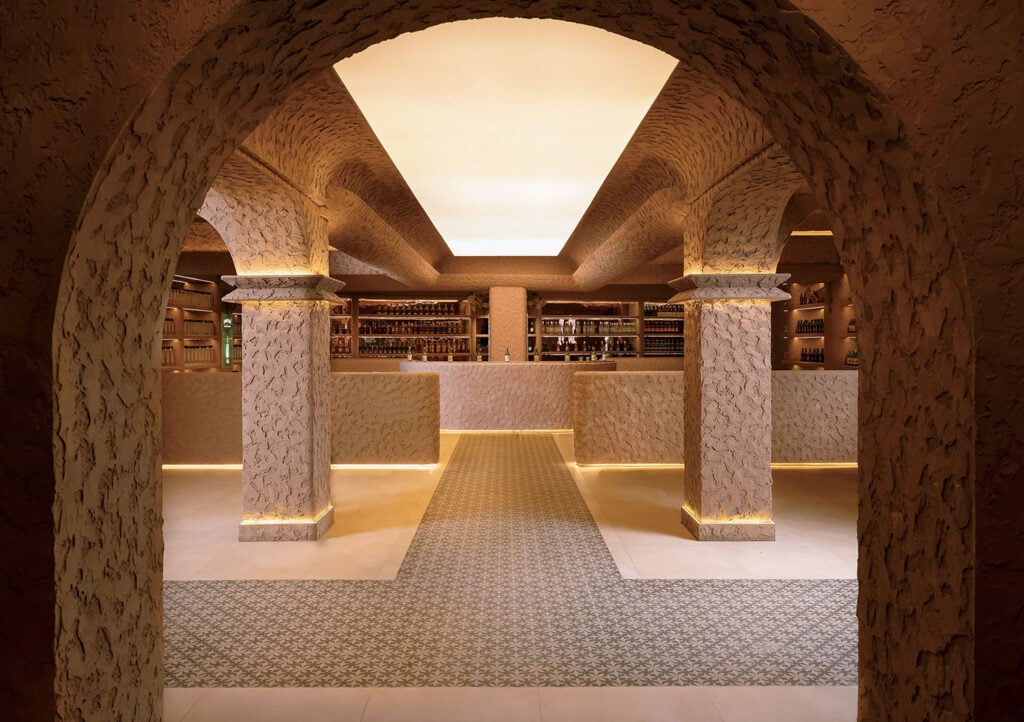 A spacious, warmly lit bar with textured stone arches, columns, patterned flooring, and shelves stocked with bottles behind a long counter.