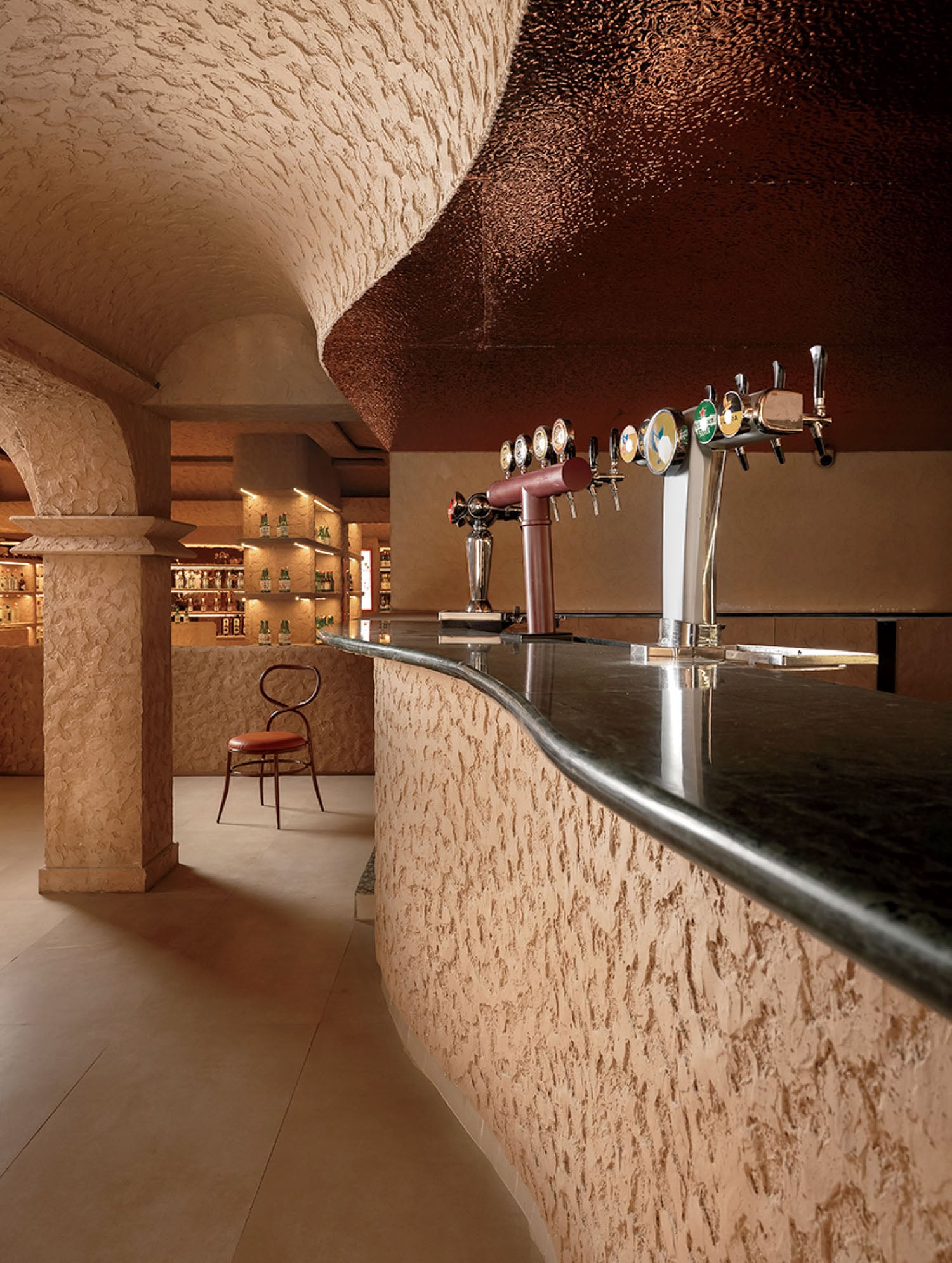A bar with a textured stone counter, multiple beer taps, arched columns, warm lighting, and a single chair in the background.