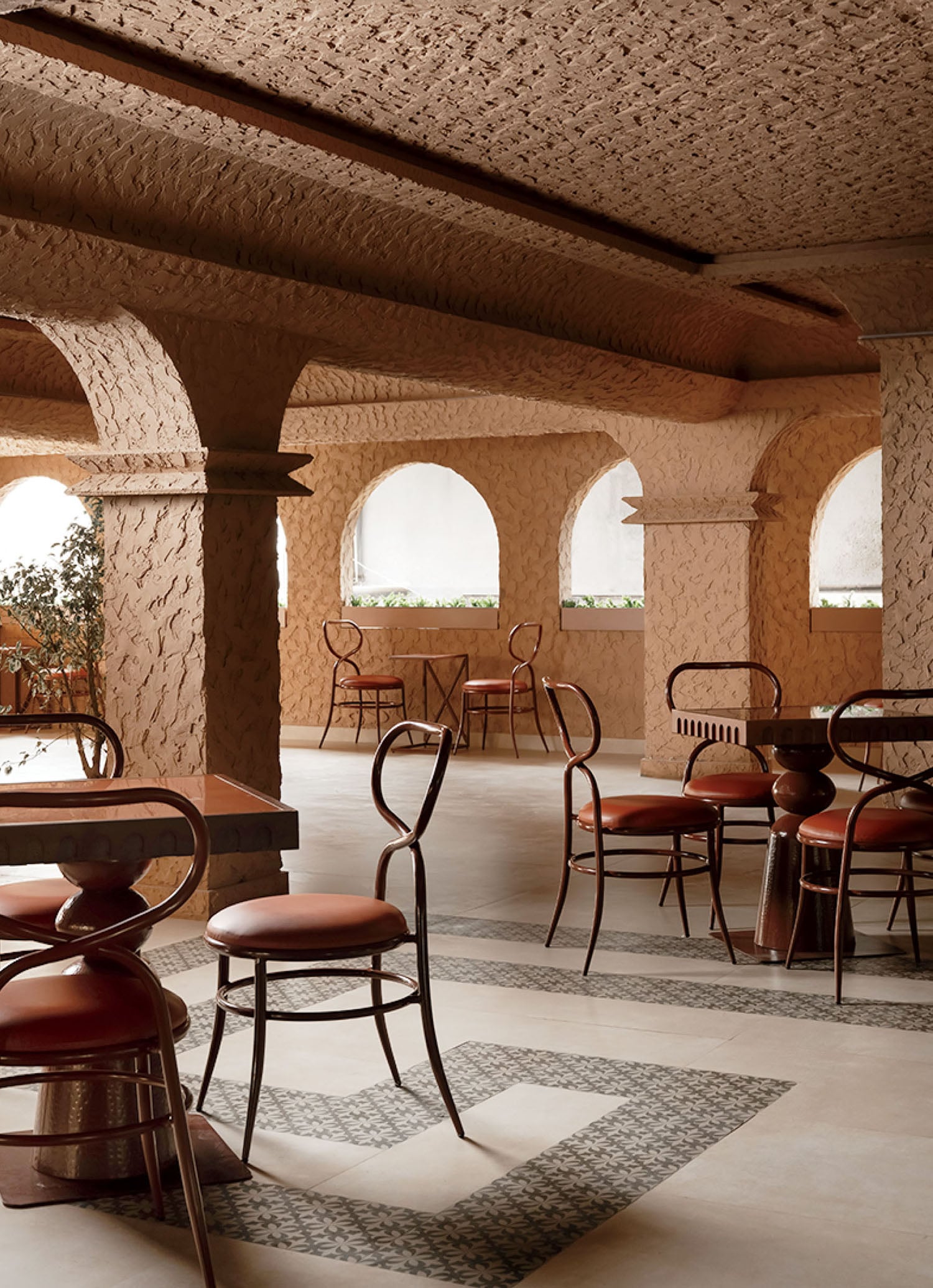 A cafe interior with textured stone walls, arched windows, metal chairs with round brown cushions, and patterned tile flooring.