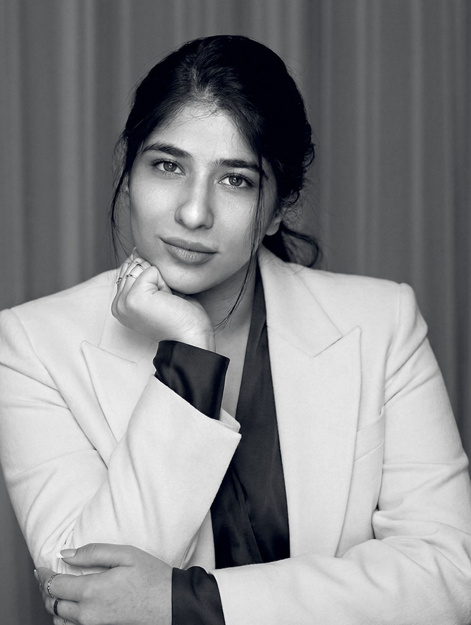 A woman with long dark hair, wearing a light-colored blazer, rests her chin on her hand and looks directly at the camera against a plain background.
